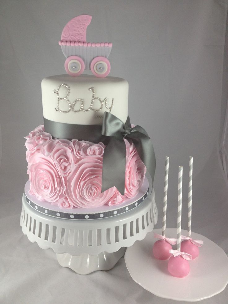 pram figurine in pale grey and pink, on top of a two-layered cake, bottom layer decorated with pink frosted roses, upper layer with the words baby written in silver, baby shower cakes for girls, shiny grey bow, and three cake pops