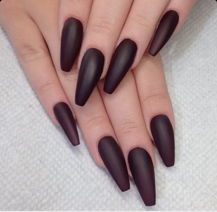 acrylic nail shapes, painted in a dark, cherry red matte nail polish, on two hands, one resting on top of the other, on a white textured surface