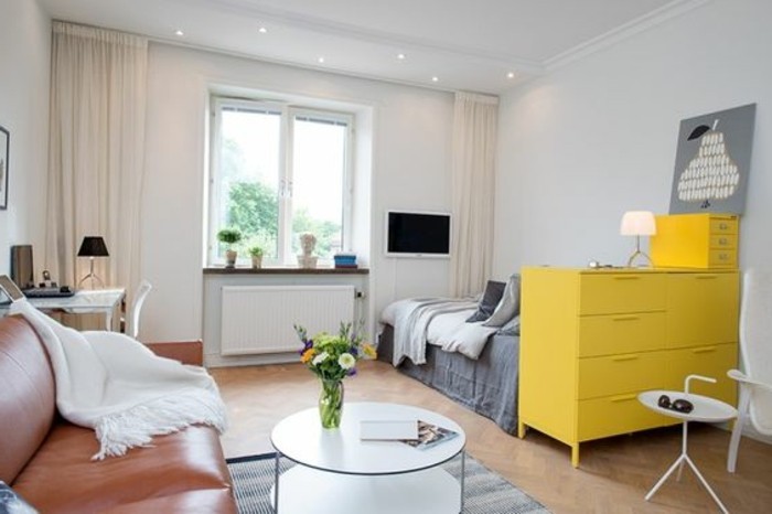 leather sofa in brown, with a white throw, near a round white coffee table, inside a room with a laminate floor, a bed and a yellow chest of drawers