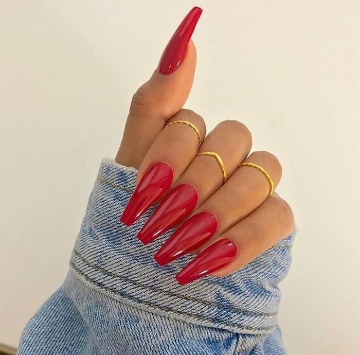 denim sleeve held by a hand, with three thin golden rings, and long and coffin nails, painted in a vibrant red color
