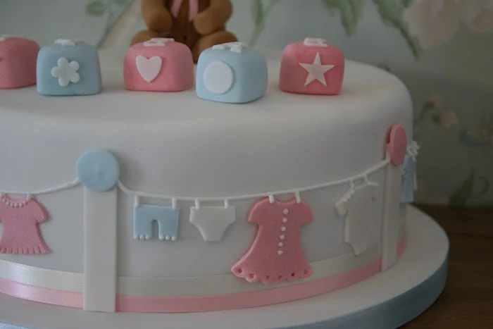 smooth white frosting, on a cake seen in close up, decorated with building blocks, in pastel pink and pale blue, with little clothes line, featuring small white blue and pink clothes, made from fondant