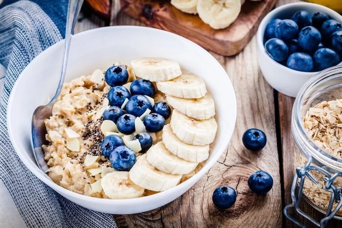 flakes of almond and coconut, on top of a creamy porridge, with sliced banana, and whole blueberries, simple breakfast ideas, ingredients in different containers nearby