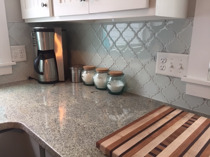 chopping board and three small jars, on a spotted beige counter top, near white cabinets, pale blue arabesque backsplash, and a coffee machine