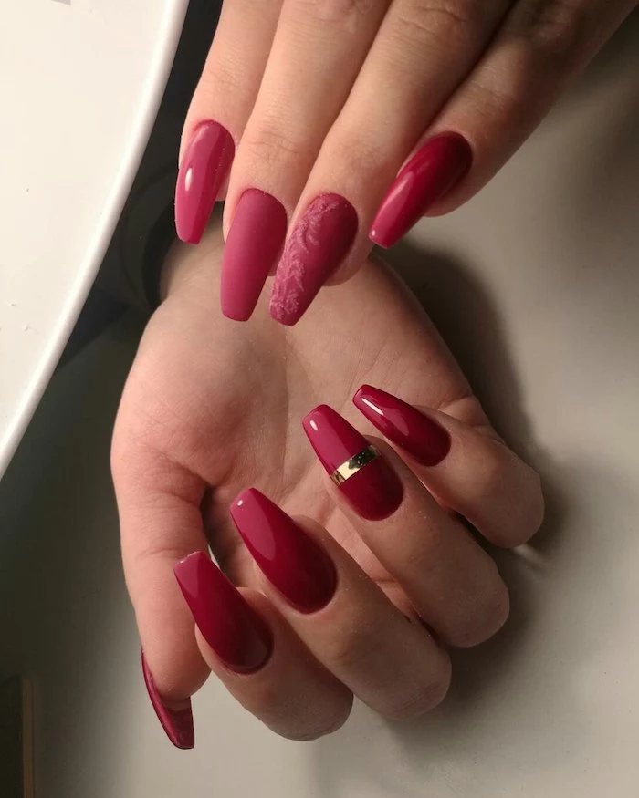 glossy and matte nail polish, in red hues, on two hands with acrylic nail shapes, one nail is decorated with a gold metallic stripe, wile another has a floral acrylic motif