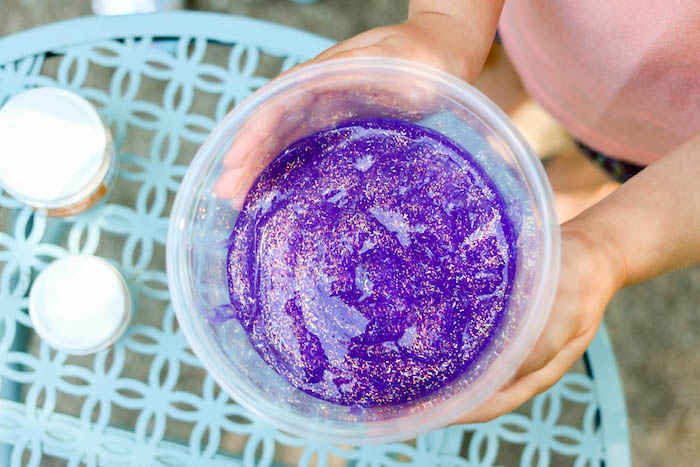 plastic clear bowl, containing dark purple goo, with light sparkling glitter, held by a child, how to make slime without borax, light blue painted wrought iron table