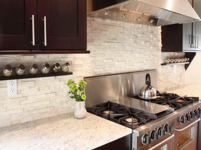 vase with flowers, a metal teapot, and several condiment jars, on brown wooden shelves, in a kitchen with an off-white, stacked stone backsplash, and brown cabinets