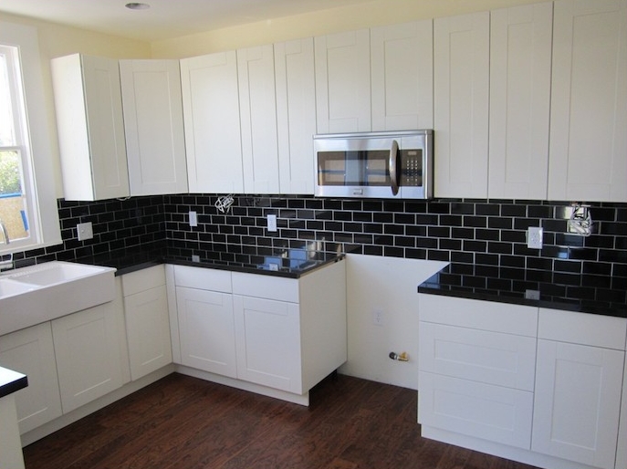 black subway tile kitchen backsplash, with white joints, inside a kitchen with pure white cabinets, dark wooden floor, and pale yellow wall