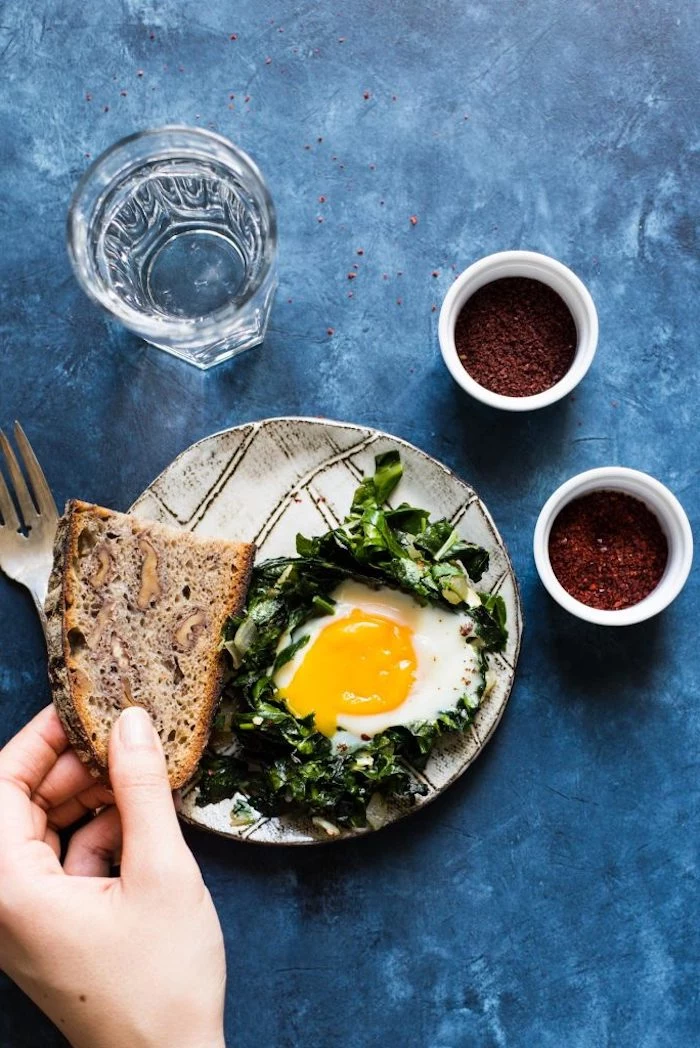hand holding a piece of full grain bread, near a plate containing spinach, and a sunny side up egg, breakfast menu ideas, glass of water and condiments