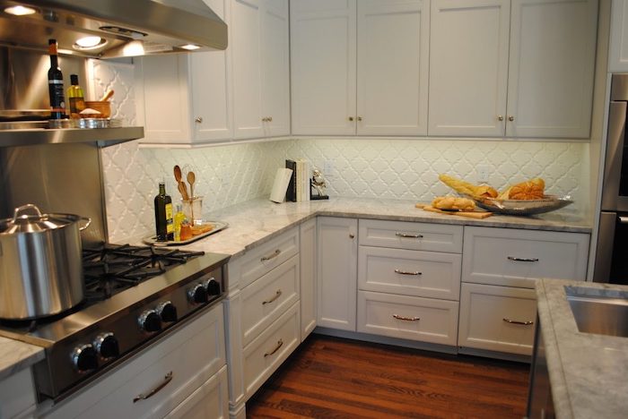 extractor hood and stove, with a large metal cooking pot, inside a kitchen, with a light blue, arabesque tile backsplash, white cabinets and a brown laminate floor 