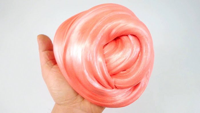 peach-colored or light coral pink, metallic fluffy slime, twisted into a roundish shape, and held by a tiny child's hand, on a pale gray background