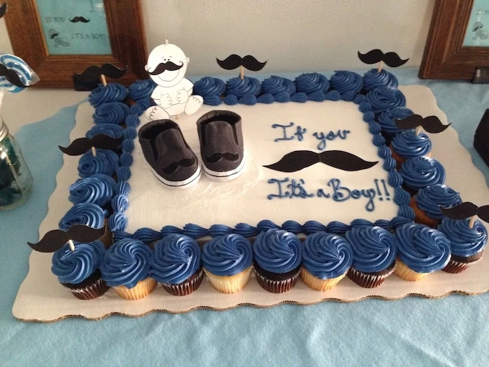 mustache toppers in black, decorating a large white cake, with small dark grey baby shoes, and a smiling baby drawing, baby shower sheet cakes, surrounded by dozens of dark blue cupcakes