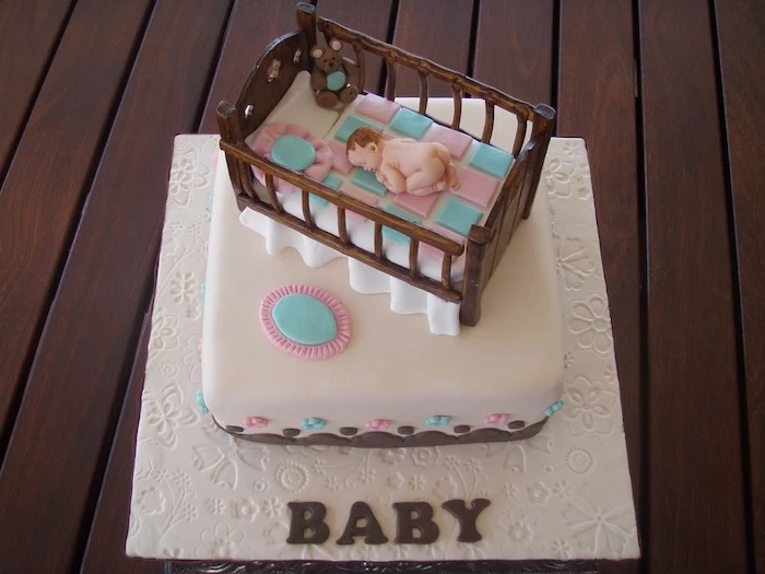 cradle made from brown fondant, containing a checkered blue and pink blanket, pillows and a sleeping baby figurine, on top of a square white cake