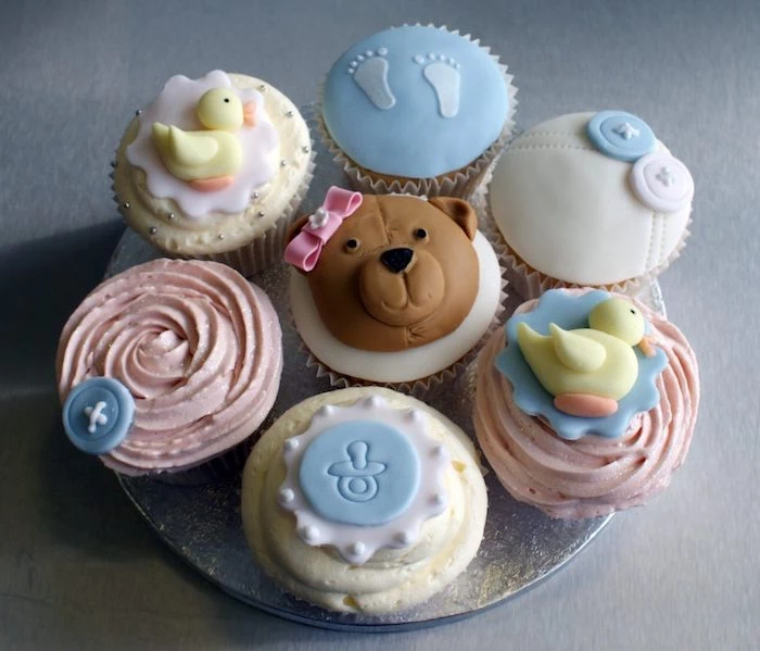 seven cupcakes with pale pink, and light yellow frosting, decorated with little fondant shapes, duckling and a teddybear, little footprints and blue and white buttons