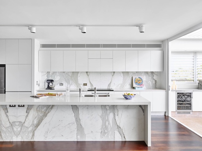 bright spacious kitchen, with smooth white cabinets, dark brown floor, white kitchen island, with a marble-effect, and a matching back splash