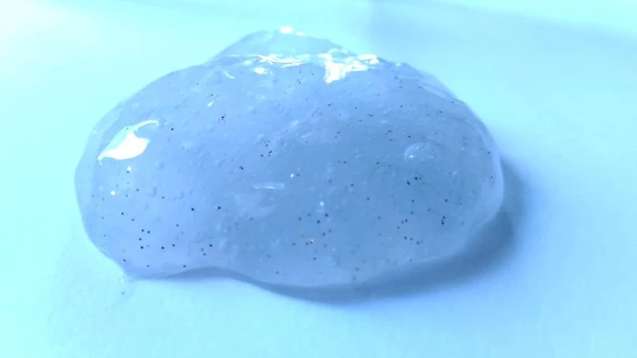 oily blob of light blue goo, decorated with fine glitter, and placed on a pale turquoise surface, slime without borax