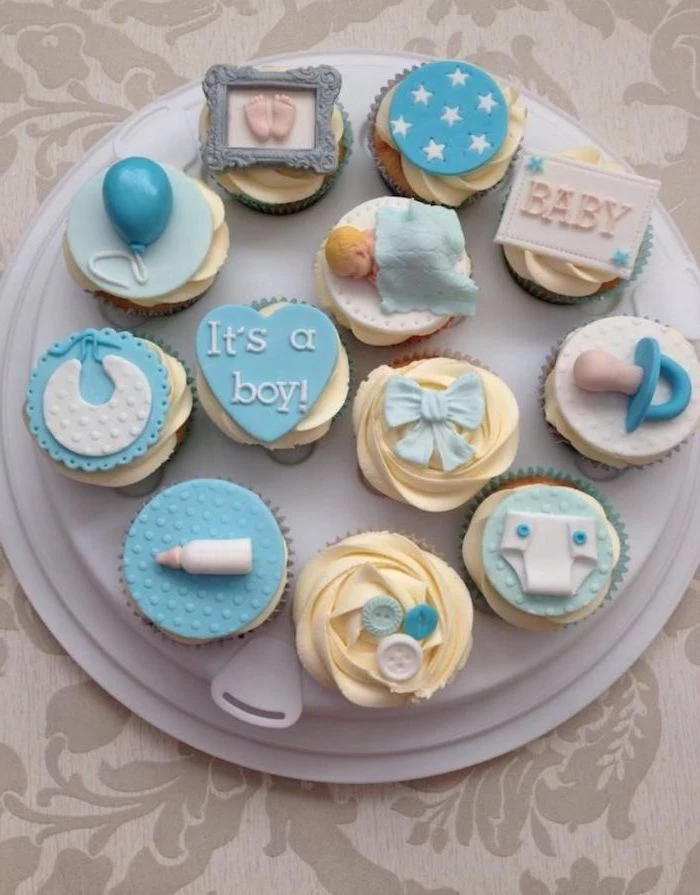 dummy and bib, baby bottle and balloon, little footprints and buttons, nappy and a sleeping baby figurine, all made from fondant, decorating a dozen of cupcakes