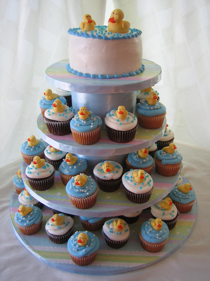cupcakes with white and blue frosting, each decorated with a small yellow ducking, made from fondant, on a cake stand, with a small round cake on top