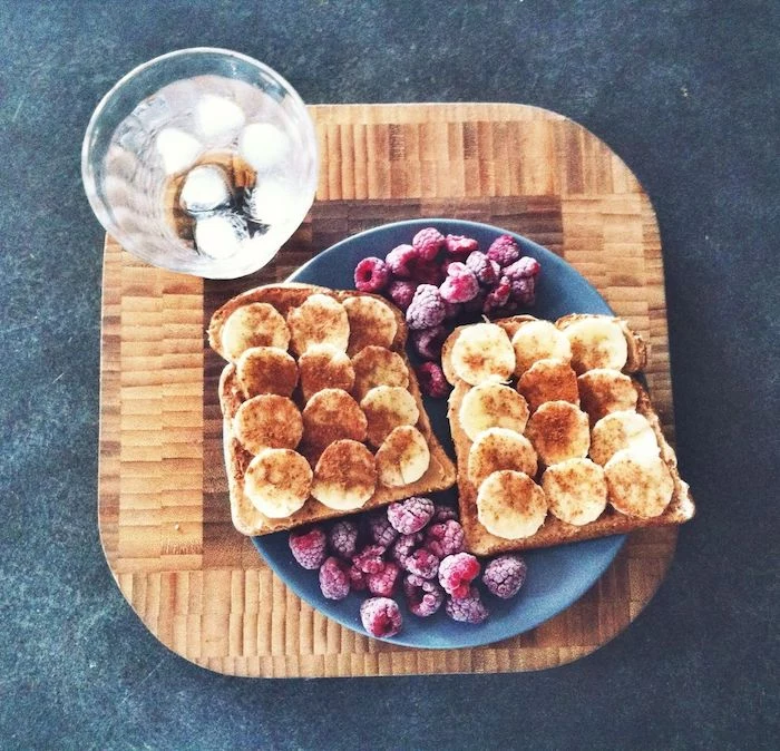 ice water in a clear glass, placed on a large wooden cutting board, near a blue round plate, with two pieces of toast, covered with banana slices and cinnamon, and accompanied with frozen raspberries, simple breakfast ideas