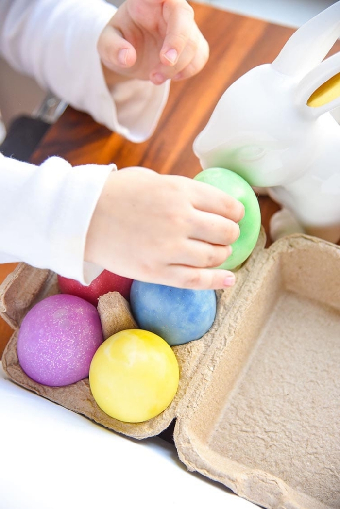 tiny hand holding a green, egg-shaped piece of slime, over an egg carton, containing red and blue, purple and yellow eggs, how to make slime with glue