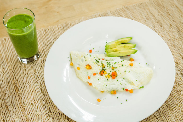 glass of green juice or a smoothie, next to a round white plate, containing an omelette, made with egg whites, topped with veggies, best breakfast for weight loss, with avocado slices on the side