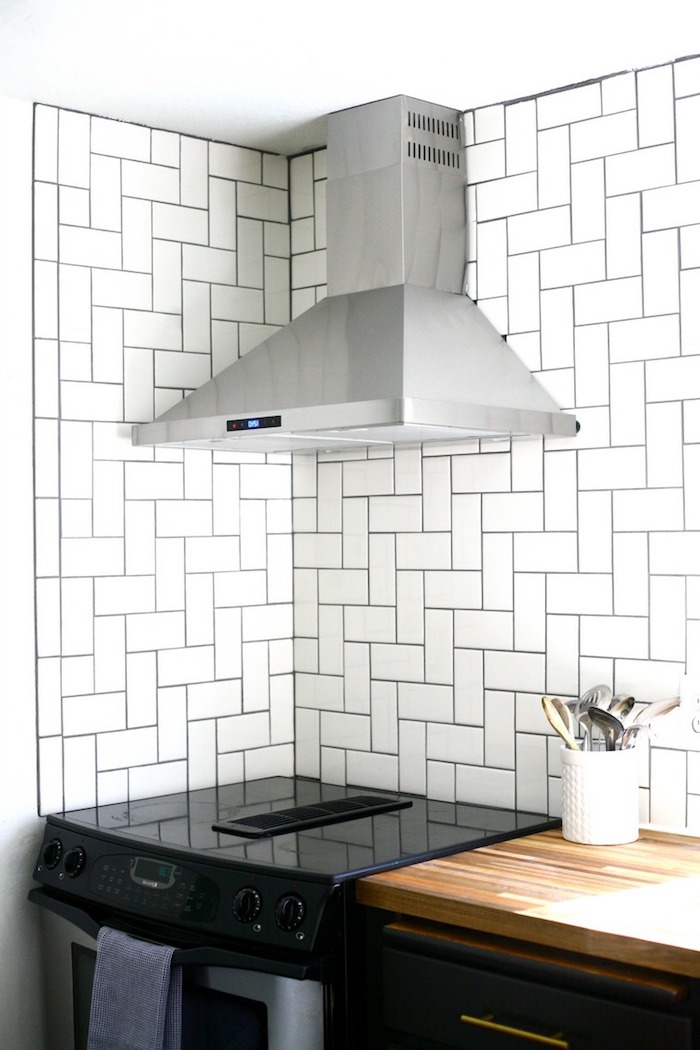 contrasting herringbone backsplash tiles, with black joints, near a glossy black oven, and a metal extractor hood