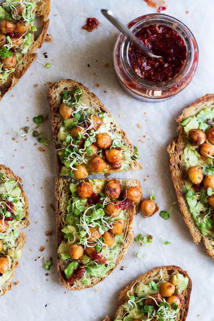 country bread slices, with guacamole and chickpeas, garnished with salad greens and sprouts, low calorie breakfast, on a pale surface, near a small jar with cranberry sauce