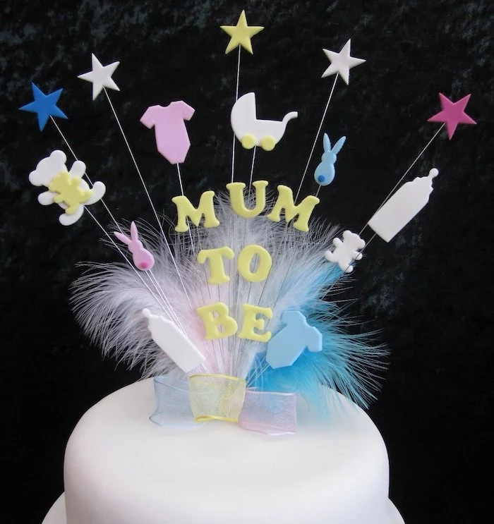 mum to be written in yellow, on a cake topper, with pale blue, yellow and pink ribbon, light colored feathers, and various baby-related shapes