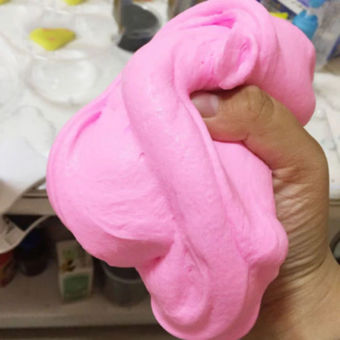 squeezing a piece of soft, pink and fluffy slime, in an adult's hand, desk with various art supplies, in the background