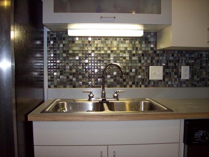 rectangular lit fluorescent lamp, near a stone and glass mosaic tile backsplash, in different shades of grey, metal sink and a light wooden counter top