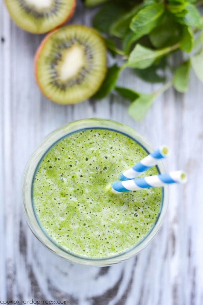 kiwi pieces and mint leaves, on a grayish wooden surface, near a clear glass, filled with foamy green liquid, healthy breakfast ideas, two paper straws