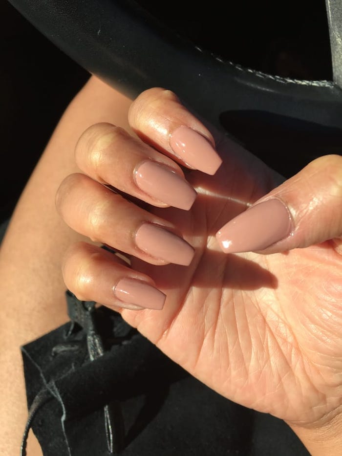 nude coffin nails, in a pinky beige hue, on a hand with folded fingers, seen in close up, near a car's steering wheel
