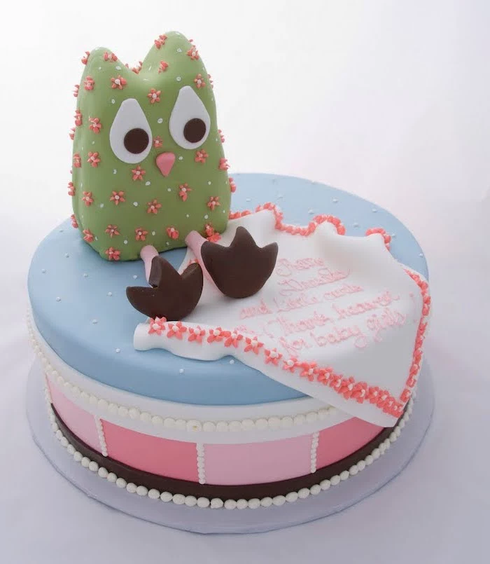 green owl figurine, with big white eyes, decorated with a pink and white floral pattern, on top of a blue and pink cake, owl baby shower cake, with a festive message, done in peach-colored frosting