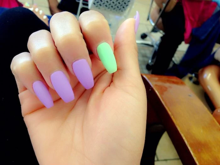 accent nail in neon green, and four other nails in neon purple, on a hand with folded fingers, matte coffin nails 