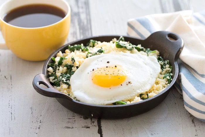cup of tea or coffee, what is a healthy breakfast, next to a pan, filled with couscous and green veggies, and topped with a large fried egg