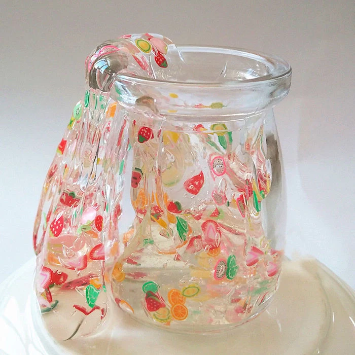 clear slime decorated with many colorful stickers, shaped like different fruit, pouring out of a clear glass jar, placed on an upturned white plate