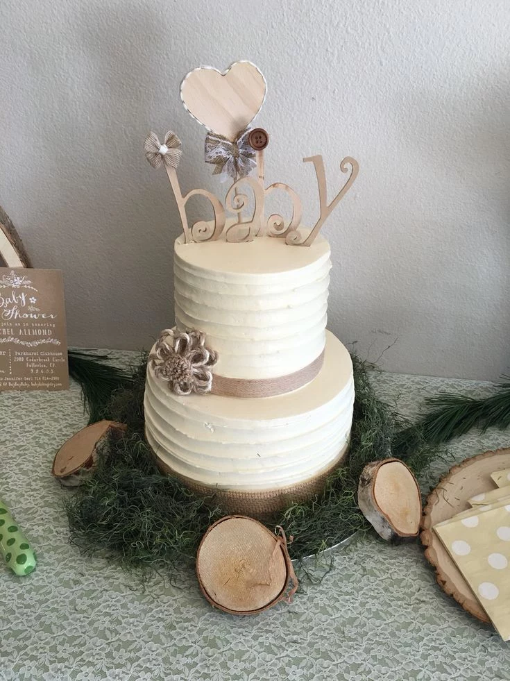 beige and cream cake, decorated with a topper reading baby, and surrounded by pine branches, and little wooden decorations