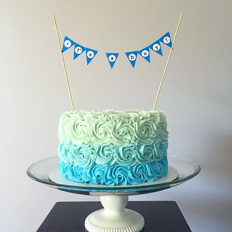 roses made from frosting, in teal and turquoise and pale minty green, on a cake decorated with tiny blue flags, spelling the words it's a boy, baby shower cakes for boys, glass cake stand