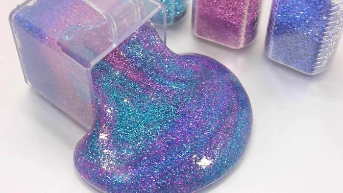 overturned clear plastic container, spilling sparkly smooth slime, in teal and purple, blue and pink, three bottles of glitter, in different colors