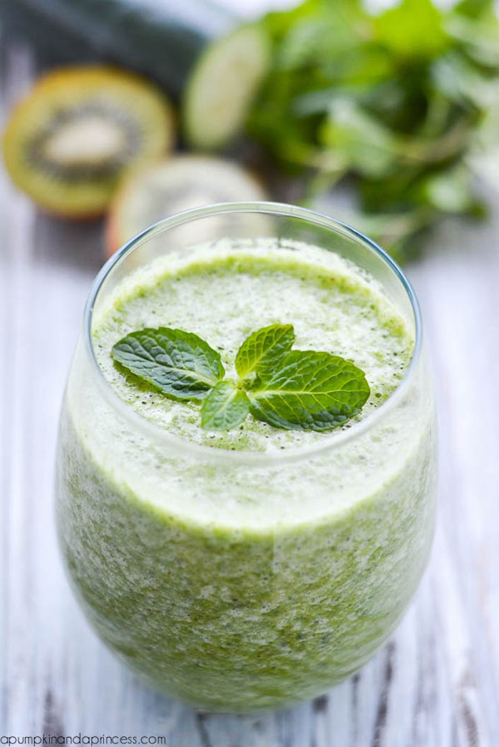 mint leaves topping a green smoothie, inside a clear tumble glass, placed on grayish wooden surface, healthy breakfast ideas, cucumber and kiwi, and mint leaves in the background