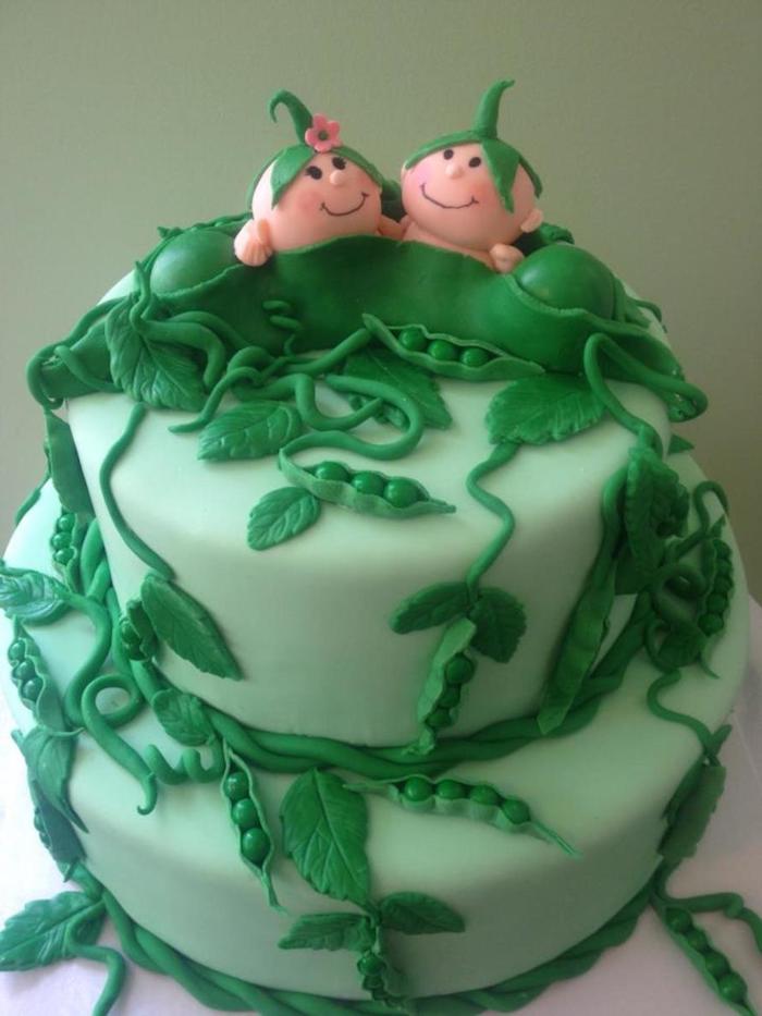 pea pod made from green fondant, containing two smiling baby figurines, and two peas, topping a light green two-layered cake, twin baby shower cakes, with pea shoots and dark green leaves 
