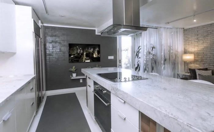 dark and light grey brick walls, in a modern kitchen, with pale grey marble countertop, fridge and cooker, potted plants and a lit lamp