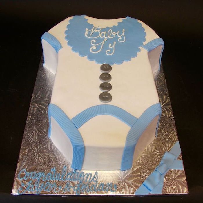 bib in blue, decorated with the words baby Jy, made from fondant, and topping a white onesie cake, with blue trims, and four grey buttons