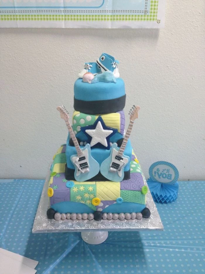 sneakers in white and blue, and a small sleeping baby figurine, in a pale blue onesie, topping a colorful patchwork cake, decorated with two blue fondant guitars