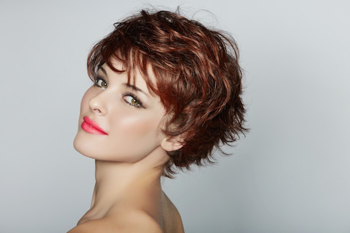 pixie cut with layers and side bangs, in auburn brown, worn by smiling young woman, with neon pink lipstick, and black mascara
