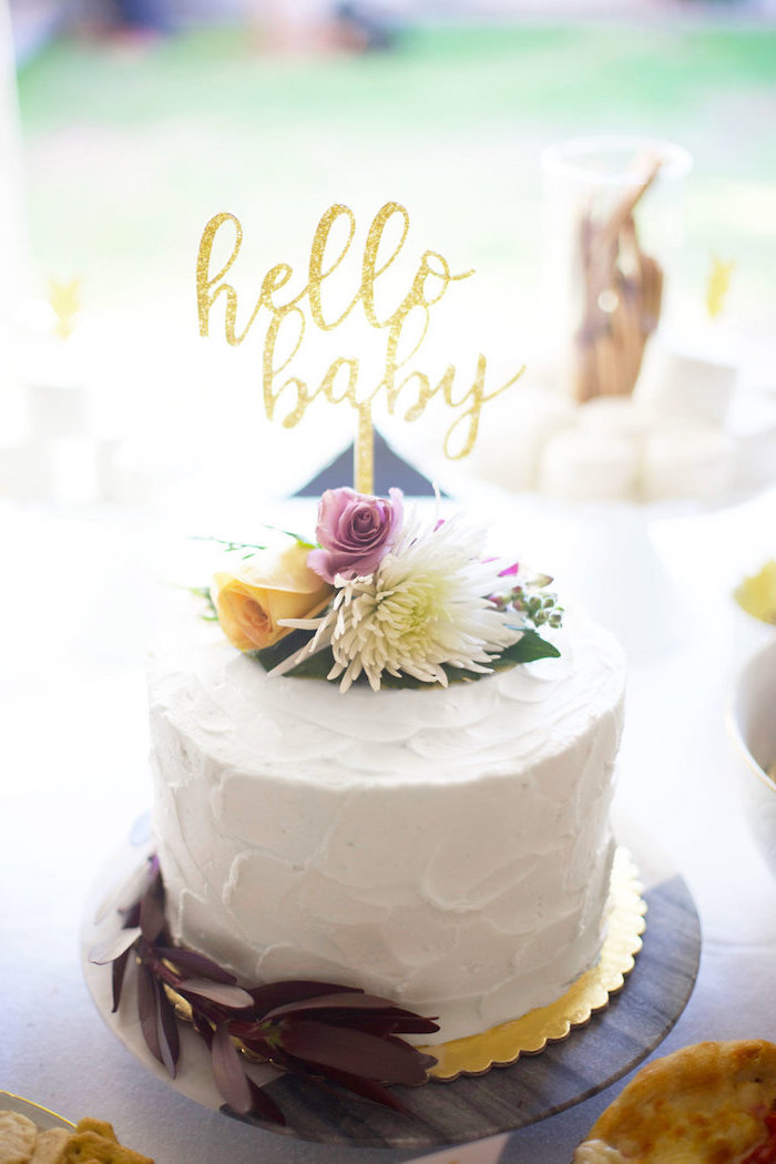 gold cake topper, with the words hello baby, on a creamy white cake, decorated with fresh flowers, in yellow and pink