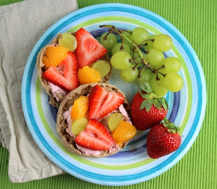 pink yoghurt spread, on two pieces of bread, garnished with chunks of strawberry, orange and grape, easy breakfast recipes, in a plate with strawberries, and more grapes