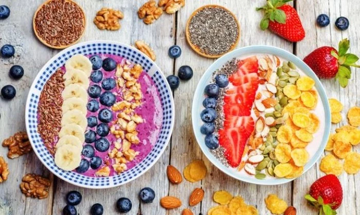 two bowls with pink and white yoghurt, topped with various seeds, cornflakes and almonds, banana slices and blueberries, strawberries and walnuts