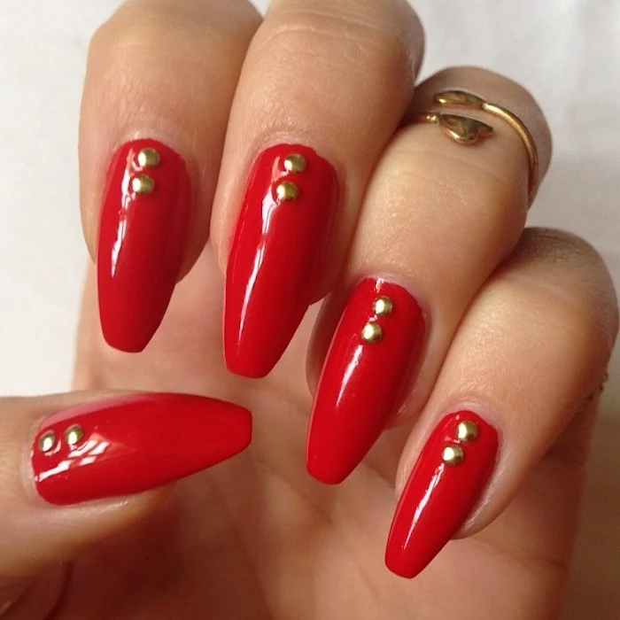 classic red nail polish, on a hand seen in close up, with coffin shaped nails, each nail is decorated with two gold rhinestones