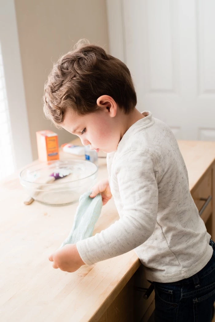 little boy stretching a piece of light blue goo, over a light wooden desk, clear glass bowl, spatula and ingredients nearby, fluffy slime recipe 