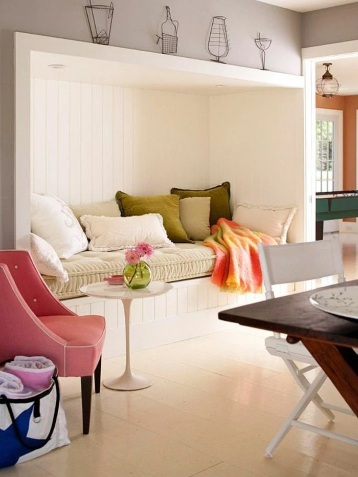 sitting nook inbuilt in a wall, with white wooden paneling, and several different pillows, in white and green, pink armchair and a small white coffee table, room design